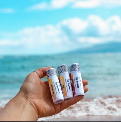 Hand holding lip butter tubes with a beach and ocean background