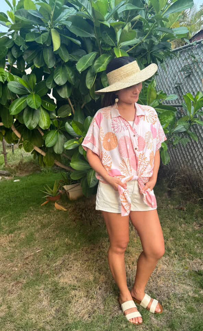 Woman in a pink floral shirt and white shorts standing outdoors with greenery in the background