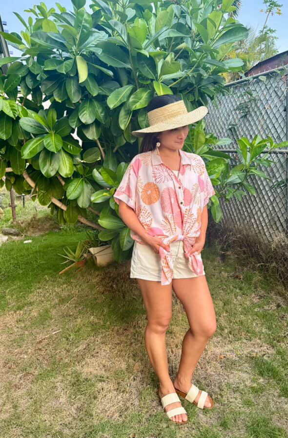 Woman in a pink floral shirt and white shorts standing outdoors with greenery around